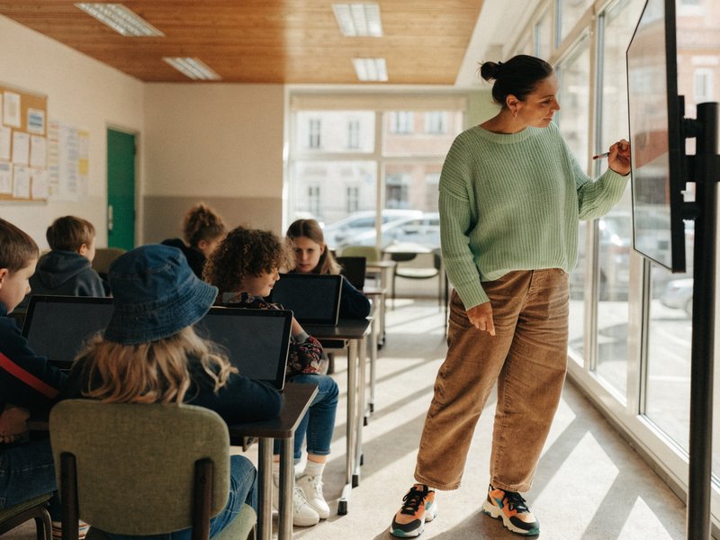 A teacher at the classroom screen, pupils at their desks with tablets.